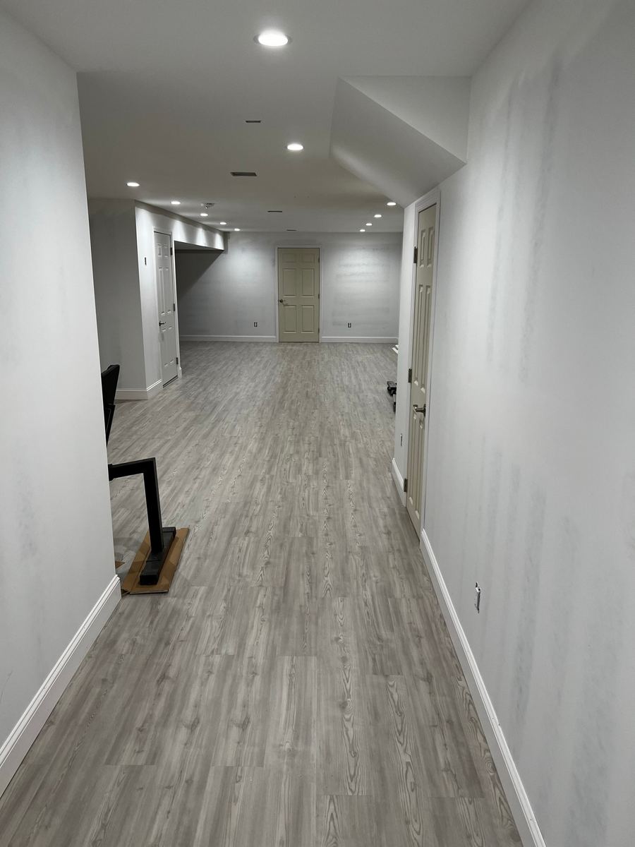 A finished basement hallway with a gray plank floor, white walls and recessed lights, the stair railing at left