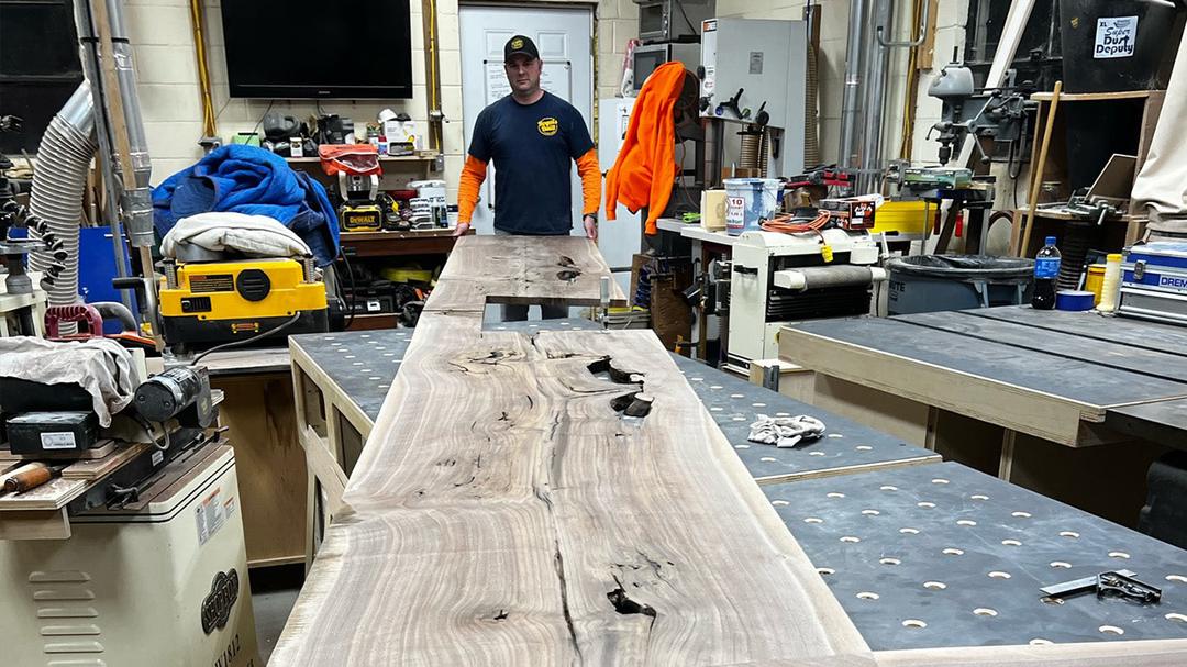 Man in a Royals Built cap standing behind a long live-edge tabletop in a busy workshop