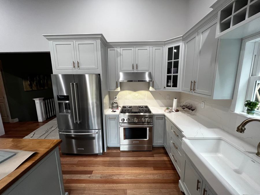 A finished kitchen: gray inset cabinets, the fridge in a tall surround, a range under its hood, a glass-front corner cabinet with divided panes, a drawer stack, an apron-front sink under a window with a wine rack above it, and a wood-topped island in the foreground