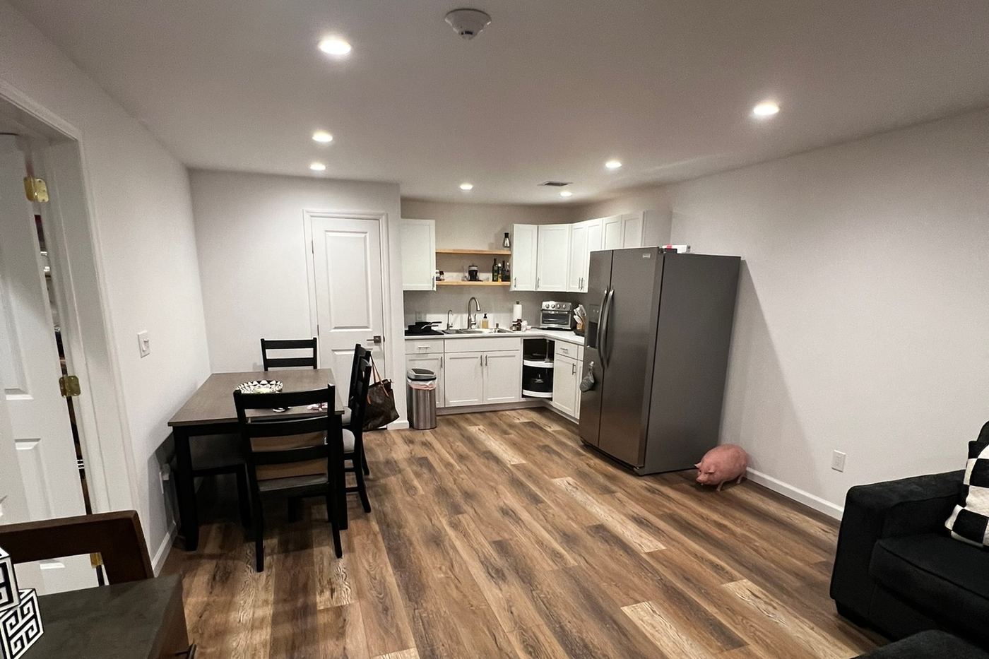 A finished basement: wood-look plank floor, white walls and recessed lights, a dining table, and a kitchenette with white cabinets and a stainless fridge at the far end