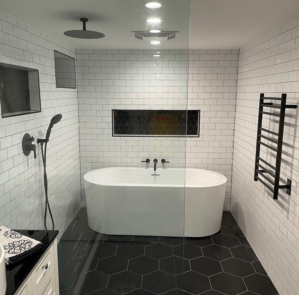 After: the same bathroom finished in white subway tile, with a rain head in the ceiling, a glass shower wall, a freestanding tub under a long black-hexagon niche, a black heated towel rack and a black hexagon tile floor