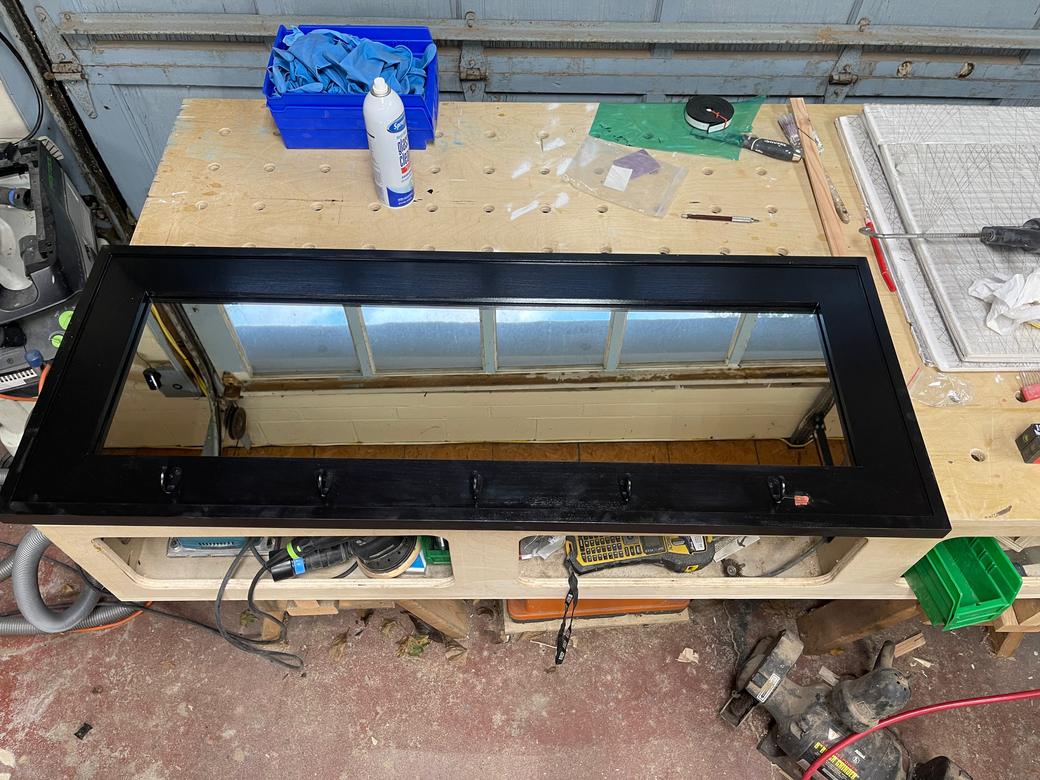 Black mirror frame with hooks along its bottom rail, lying on a workbench in the shop