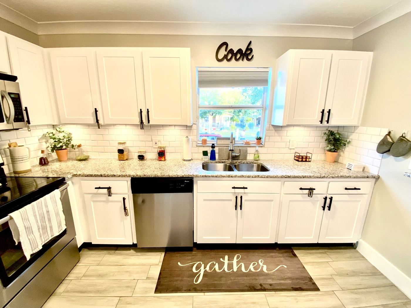 White shaker kitchen with black bar pulls and crown molding, a white subway-tile backsplash, light granite counters and a stainless sink under a window