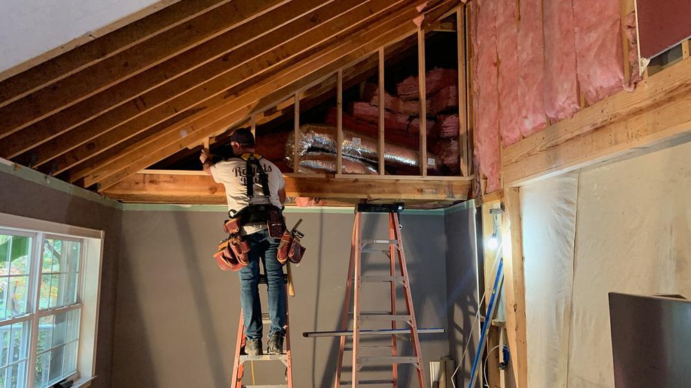 Worker on a ladder framing a cathedral ceiling, seen from behind, with the Royals Built logo on his shirt