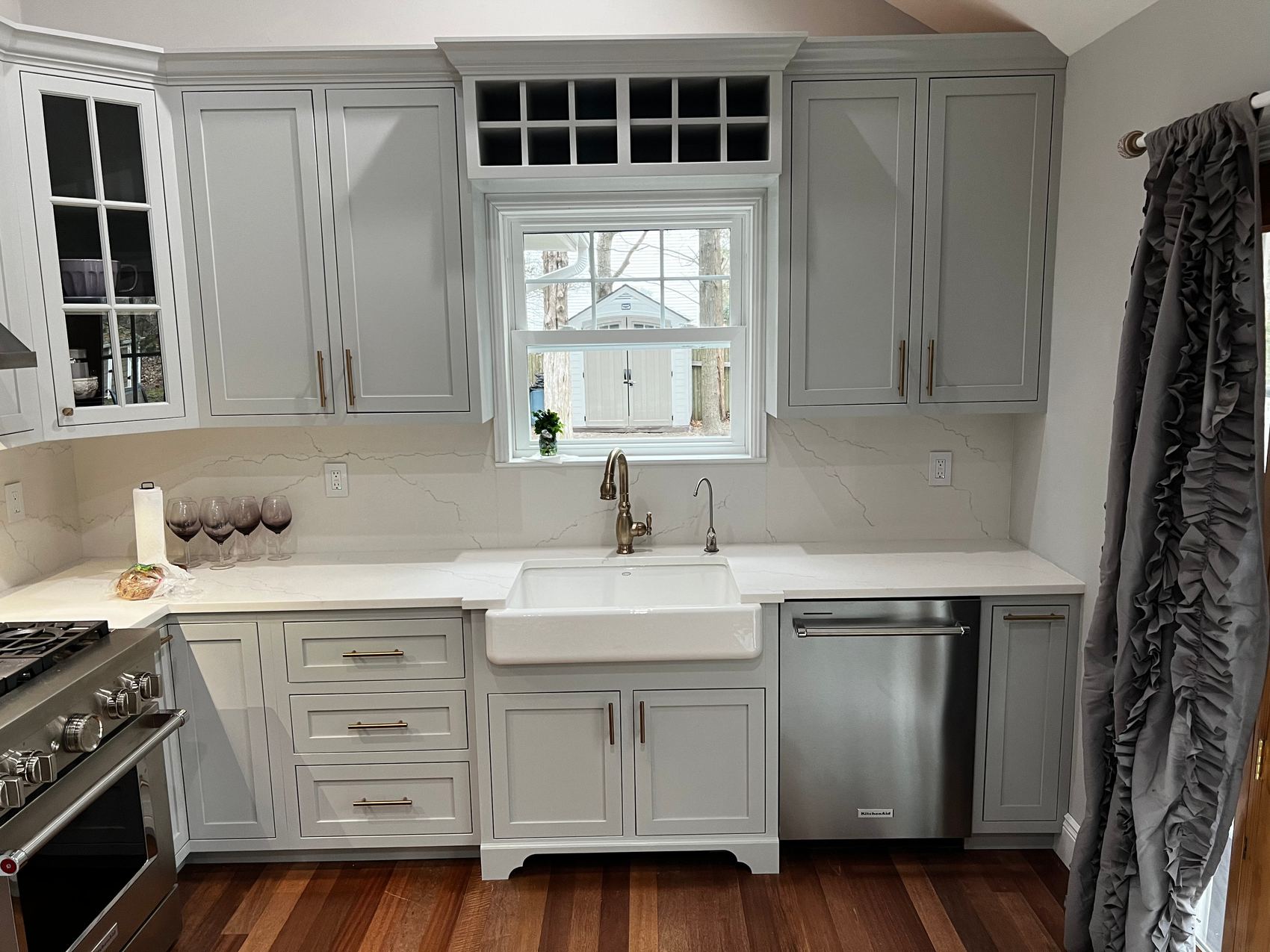 Sink wall of a kitchen with gray inset cabinets, a glass-front upper, a wine rack above a window and a white apron-front sink